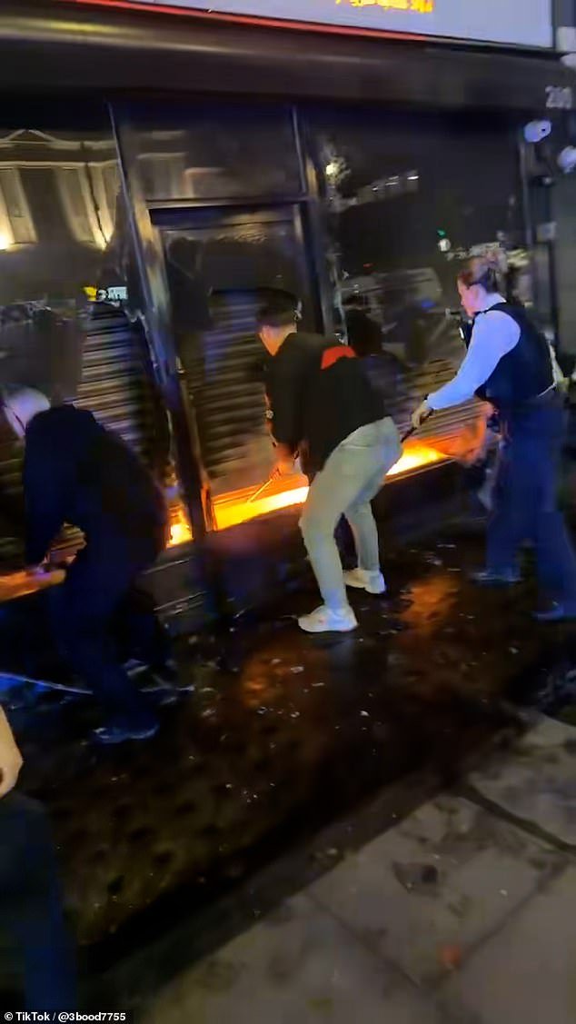 Los transeúntes del héroe del momento entran a la fuerza en una tienda de comestibles en llamas para salvar al hombre atrapado en su interior, mientras rompen ventanas y rompen el cierre de seguridad.